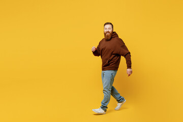 Full body smiling happy fun cheerful satisfied young redhead caucasian man wearing brown hoody casual clothes walk go look camera isolated on plain yellow background studio portrait Lifestyle concept