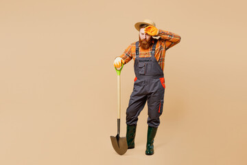Full body tired sad young bearded man wears straw hat overalls work in garden hold shovel digging wipe sweat isolated on plain pastel light beige color background studio portrait Plant caring concept