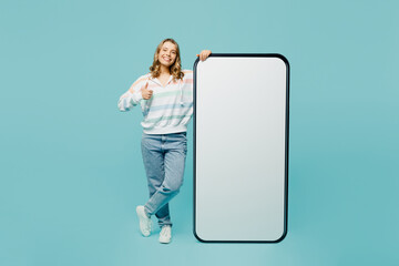 Full body young fun woman wear striped hoody big huge blank screen mobile cell phone smartphone with workspace copy space mockup area show thumb up isolated on plain pastel light blue cyan background.