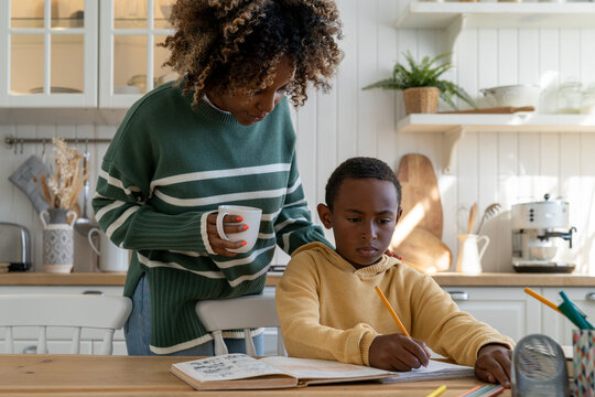 African American mother checking childs homework, mom parent supporting kid with learning, young woman mom standing over schoolboy helping son to succeed at school. Parental involvement in education