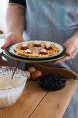  A lady in a blue striped apron holds a plate with a waffle garnished with strawberry jam and cream.