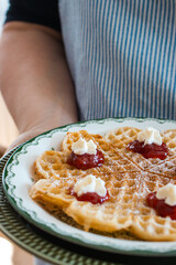 A lady in a blue striped apron holds a plate with a waffle garnished with strawberry jam and cream.