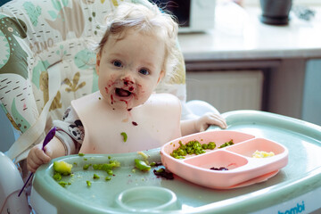 Cute baby girl tries to eat her first meal. Baby-Led Weaning (BLW). Adorable infant eating in the...