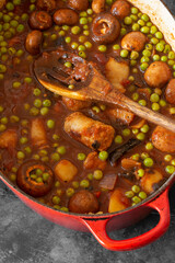 Sausage casserole in a red casserole dish, with potato, mushrooms, tomatoes, peas and onion, in a beef stock.