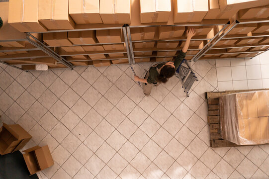 Top View Of Employee Preparing Customer Order In Storehouse, Checking Cardboard Boxes. Stockroom Worker Wearing Industrial Overall Working At Packages In Storage Room. Warehouse Concept