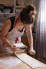 A beautiful Italian Woman rolls out the dough with the help of a pasta machine, preparing a Delicious dinner at Home from flour and Kitchen utensils. The concept of technology in everyday life.