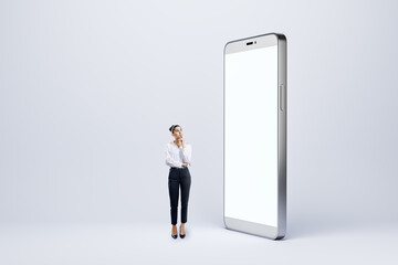 Thoughtful young businesswoman looking at big empty white smartphone on light background. Technology, innovation and communication concept. Mock up.