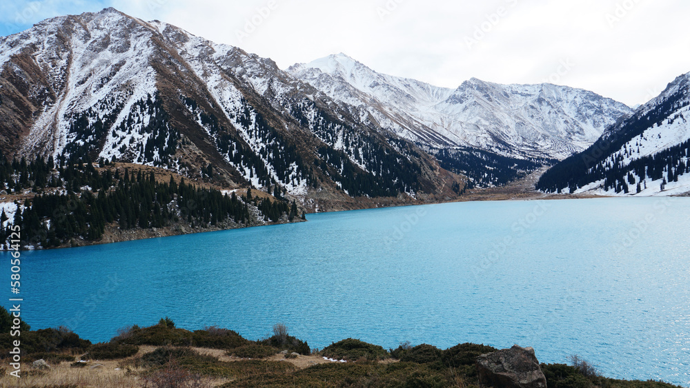 Wall mural A mountain lake with blue water in winter. The sun's rays are reflected from the water. Green forest and grass grow on the hills. There are stones lying. The high peaks are covered with snow. Almaty