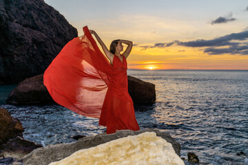 Woman in a red flying dress on the ocean or on the sea beach against the backdrop of the sunset sky.