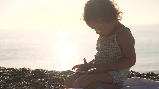 Focused baby girl uses smartphone on the beach. Gadget addiction concept