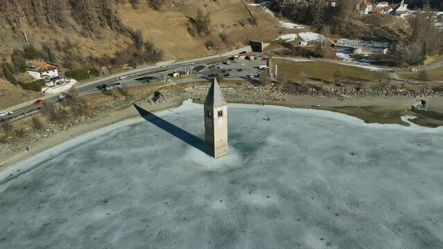 Tourists walking to the sunken church of Graun at the Reschen lake on a cold winter day. Drone panning tilt shot