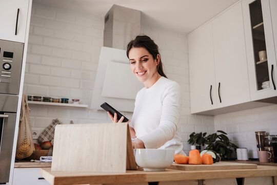 Happy Woman Using Tablet PC Holding Phone In Kitchen