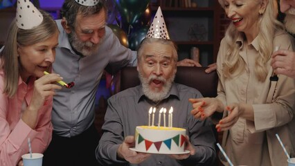 Elderly man in birthday hat blowing out candle on birthday cake, celebrating together with other senior people at retirement home. Birthday party. Spending time together, blowing party horns
