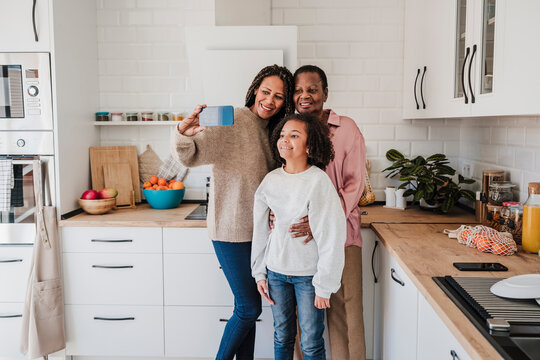 Woman taking selfie with mother and daughter through smart phone in kitchen at home - Powered by Adobe