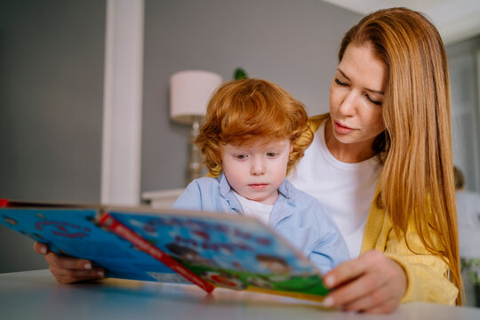 Redhead woman assisting son reading book at home