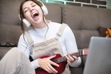 Cheerful young woman playing the ukulele, sitting in the apartment. Joyful girl holding an acoustic ukulele and singing at home. women's musical hobby, playing the ukulele