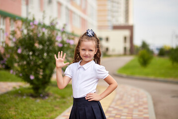 Lovely smiling first grader girl in school uniform shows hi five gesture near the school building. Happy little schoolgirl is ready to go to class for the first time.