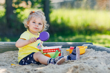Happy toddler girl playing in sand on outdoor playground. Baby having fun on sunny warm summer...