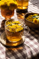Dandelion flower healthy tea in glass teapot and glass cup on table. Delicious herbal Hot tea from fresh dandelion flowers at home at summer day. Green clearing. Bouquet of dandelions petals