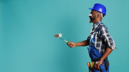 Industrial worker in uniform painting walls with roller, using renovation instrument over blue backdrop. Professional craftsman with hardhat working with rolling paint brush to change color. - Powered by Adobe