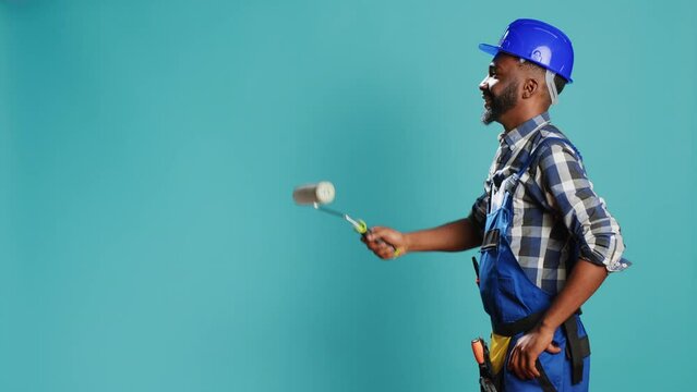 Industrial worker in uniform painting walls with roller, using renovation instrument over blue backdrop. Professional craftsman with hardhat working with rolling paint brush to change color.