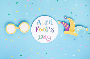 April Fool's Day holiday card with Jester hat, confetti and funny glasses on blue background, top view.