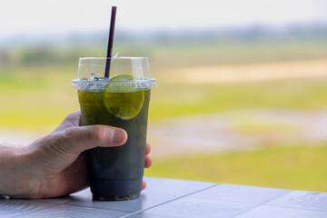 Selective focus of a plastic glass of ice green tea served with sliced lime, Hand holding a takeaways homemade cold drink lemon matcha on wooden table with blurred green garden background.