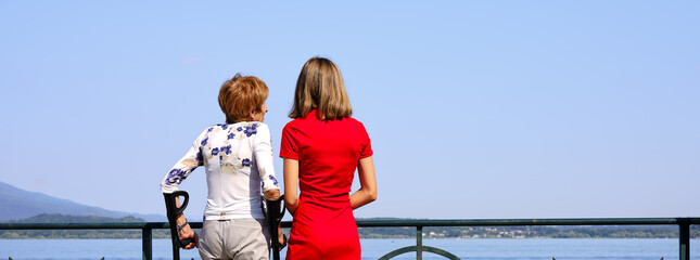 Young caregiver with elderly woman who uses crutches because she can't walk due to walking problems. Walk on the shore of Lake Maggiore in Italy.