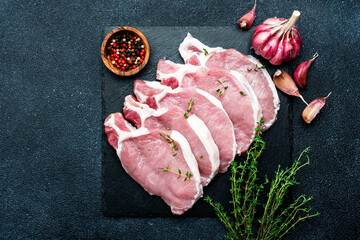 Raw pork chops on black slate board prepared for cooking with garlic, thyme, spices and pepper. Dark kitchen table,  top view