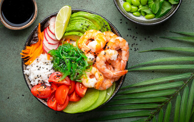 Poke bowl with shrimp, avocado, radish, carrot, tomato, chuka seaweed and white rice. Soy sauce, lime and sesame dressing. Green table background, top view