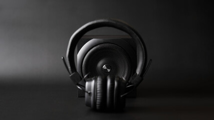 Black one-way speaker is visible through black headphones with ear pads on a black background....
