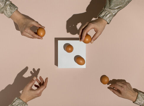 Four female hands reaching for fresh brown chicken eggs on white rectangle stand.  Minimal food concept. Flat lay.
