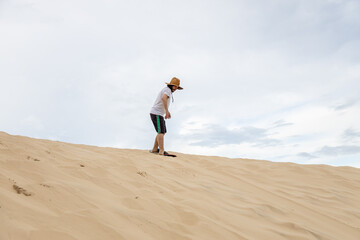 Man riding a sand board wearing a straw hat, shorts and white shirt on a sand dune in Balneario Atlantico beach, Arroio do Sal, RS, Brazil