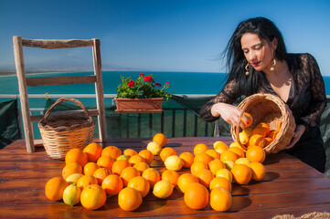 View of a typical mediterranean woman handling oranges and lemons in a panoramic balcony in Sicily, with blue sea and Mount Etna in the background - 580621768