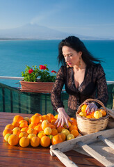 View of a typical mediterranean woman handling oranges and lemons in a panoramic balcony in Sicily, with blue sea and Mount Etna in the background - 580621797