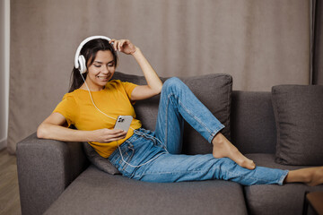 Fototapeta premium Happy indian or arabian girl resting on cozy sofa at home, listening to music