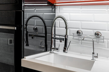 Kitchen faucet is black, near the wall with glossy white bricks and glossy black and red cabinets