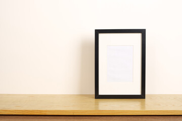 Front view of an empty black photo frame on wooden book shelf. Blank paper wall with copy space.