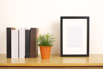 Wooden shelf with vertical black empty photo frame. Books, plant and empty background with copy space.