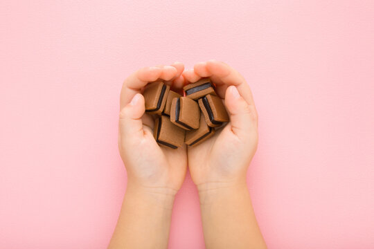 Brown Jelly Gummy Candies With Dark Black Chocolate In Baby Girl Opened Palms On Light Pink Table Background. Pastel Color. Sweet Snack. Closeup. Point Of View Shot. Top Down View.