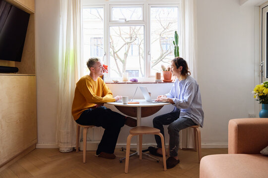 Same Sex Male Couple In Lounge Sitting At Table With Laptop Working From Home Together