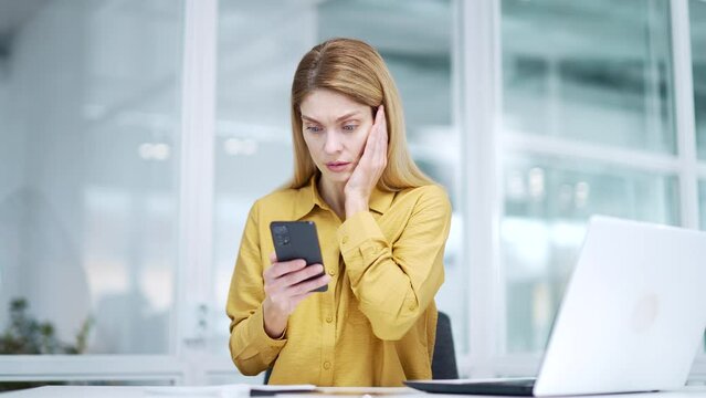 Stressed blond mature business woman entrepreneur reading message on smartphone with bad news project failure bankruptcy personal or working problems feeling shocked confused at light office workplace