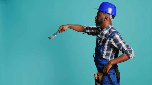Young carpenter painting walls with paintbrush on camera, using renovating tools to change color. Professional craftsman working with brush on reconstruction equipment on blue backdrop.