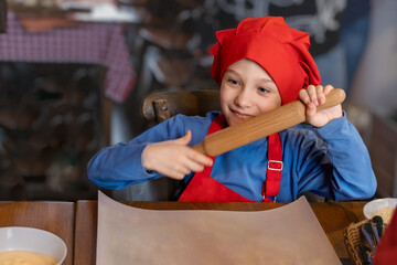 A smiling boy in the form of a chef, holding a wooden rolling pin, getting ready to cook pizza. Tools and equipment for cooking. Place for text, mockup.