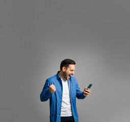 Charming mid adult man in denim shirt shouting ecstatically and pumping fist while reading good news over smart phone isolated on gray background
