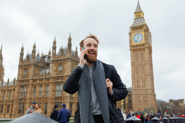Happy bearded man talking on the phone while standing on the background of the Big Ben tower - obrazy, fototapety, plakaty