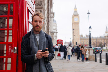 Man with a smartphone in his hands standing near a red telephone booth against the backdrop of the...