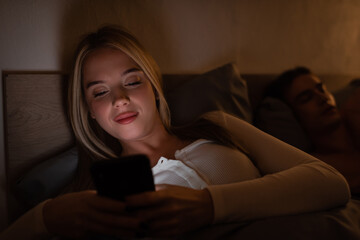 cheerful woman messaging on smartphone next to sleeping boyfriend at night.