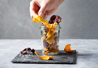 Healthy vegetable chips in a glass jar on stone cutting board with woman hand taking one of the...