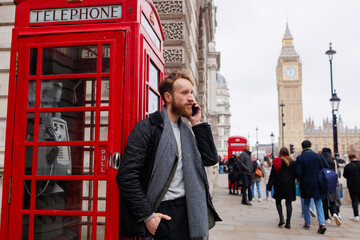 Man is talking on a smartphone while standing on a London street near a red telephone booth against...